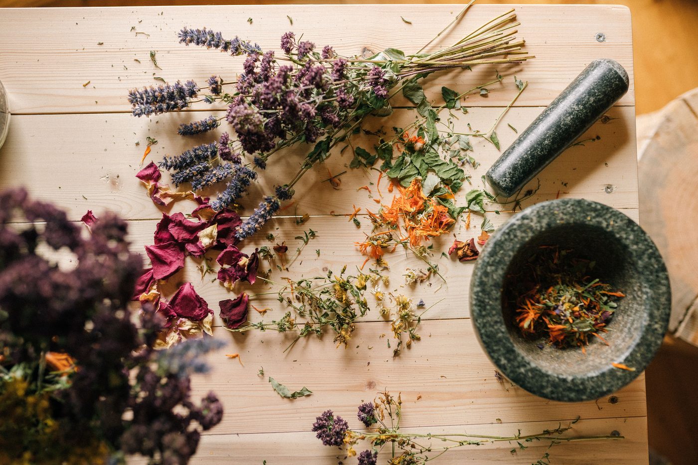 Dried medicinal herbs — lavender, calendula, and rose petals — with a stone mortar and pestle on a wooden table